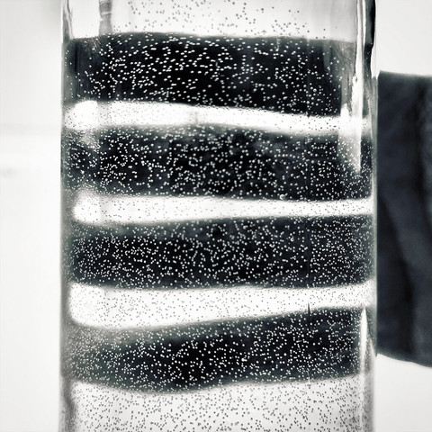 Close-up of a glass bottle filled with water, showing bubbles against dark shadows of fingers in the background.