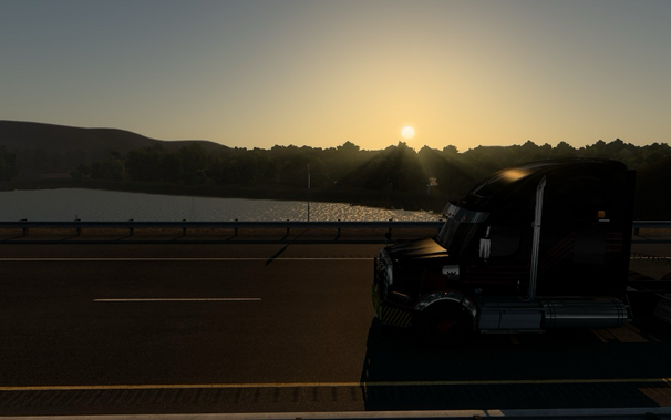 A black semi-truck is parked on the side of a highway during sunset. The sun is low in the sky, casting a warm glow over the landscape. In the background, there is a body of water surrounded by trees and hills. The scene is tranquil and picturesque, highlighting the beauty of nature and the peacefulness of the evening.