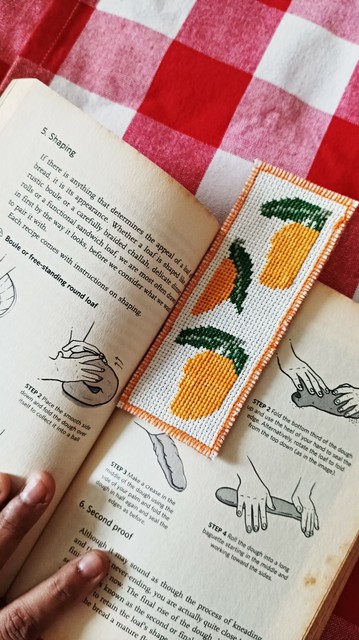 A cross stitched bookmark with mango design, inside a book about shaping doigh (for bread). The book is on a checkered table cloth, and I'm holding to keep the book stay open.