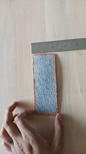 The backside of the cross stitch showing the grey backing, next to a scale and my hand.