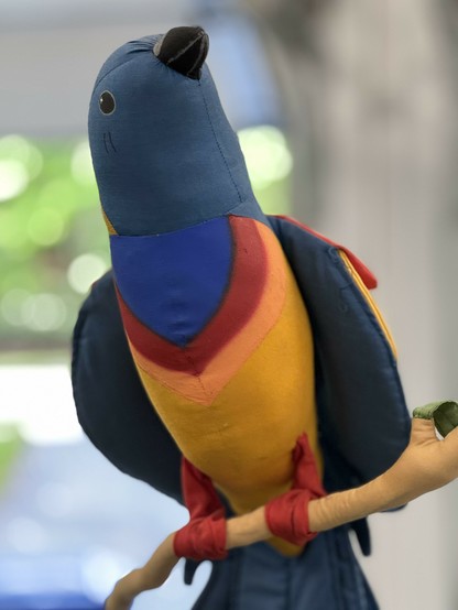 A stuffed blue-headed bird with a yellow body and a red band around his neck is perched on a "stick" which is hanging from the ceiling of my garage, although you can't see that is how he is supported.  His red fabric feet wrap around the fabric stick, which also has a green fabric leaf.  He has his neck turned, bright back eyes and a black beak.  He's very contented.