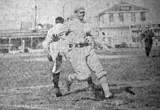 1910s black & white newsprint photo of a baseball player running with one foot landing on a base.  2 story wooden buildings are seen in the background.