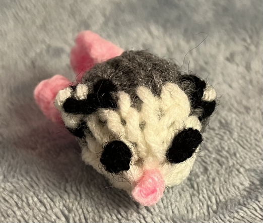 A tiny little baby opossum knitted out of gray, white, black and pink yarn. Her tail is curly and her ears are tipped with white. She has a little pink knot for a nose.
