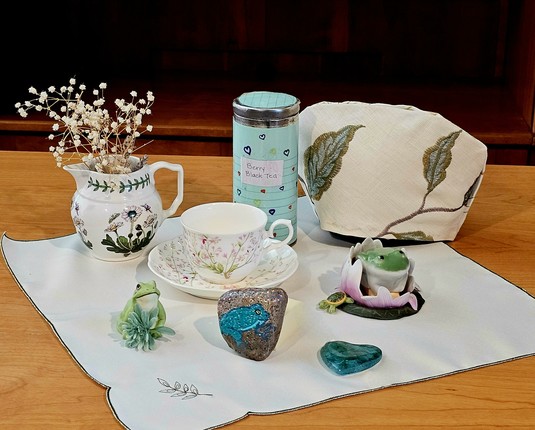 Tea tableau in white and green with a frog theme. There are a ceramic frog on a lily pad, a bisque porcelain frog with a sprig of greenery, and a rock with a frog painted on it, as well as vintage cup and saucer, a Portmeirion cream pitcher, an embroidered tea cozy, and embroidered satin tea cloth. The tea selection is Berry black loose leaf tea. There is also a flat, translucent, green carved stone in a heart shape.