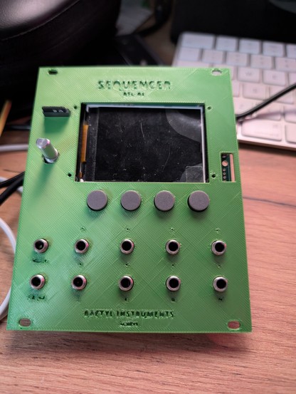 Green 3D printed Eurorack module panel, placed on top of the corresponding module (one encoder, ten jacks, a large screen and an SD card reader). There are letters carved out at the bottom: "BAETYL INSTRUMENTS" and "GENÈVE". At the very top, the name of the module, which is "SEQUENCER", subtitle "BTL-01". The module is sitting on a wooden table. It looks kind of naked (no encoder knob, no jack nuts.