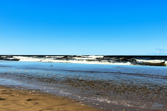 Im Vordergrund seht ihr ein Stückchen vom Ostseestrand, danach seichtes Wasser, nach der ersten Sandbank im Wasser seht ihr große Wellen. Spaß für Wasserratten. Ansonsten geht das dunkelblaue Wasser in einen hellblauen Himmel über.