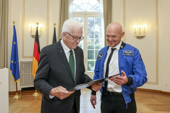 Ministerpräsident Kretschmann (links) mit Anzug und grüner Krawatte und Alexander Gerst (rechts) in blauer ESA-Jacke, betrachten gemeinsam eine Mappe in einem festlichen Raum,  im Hintergrund EU-, Deutschland- und Baden-Württemberg-Flagge. © Staatsministerium Baden-Württemberg/ Uli Regenscheit
