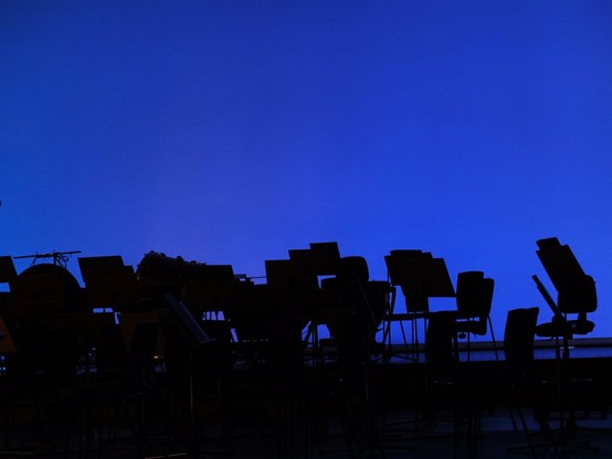 This image shows dark silhouettes of what appear to be music stands set against a solid deep blue background. The stands are arranged in a row along the bottom portion of the frame, creating an irregular jagged outline against the blue backdrop. The entire upper portion of the image consists of the uniform blue color with no clouds or other features visible. The stands are completely black, showing only their distinctive shapes with no visible details within the silhouettes.