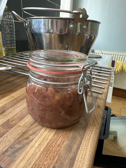 A mason style glass jar with a darkish chutney inside sits on a kitchen counter. There is a large stainless steel jam pan sits on the cooker behind.