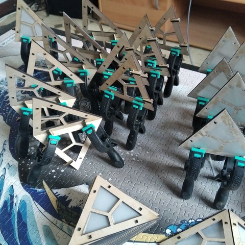 A picture showing  30 laser cut wood isosceles triangles in different configurations (some alone, some glued together on their short side).
All configurations have 2 small black and green clamps on one of their short side.
In the foreground, a stack of triangles are waiting the other to dry to get their clamps :)