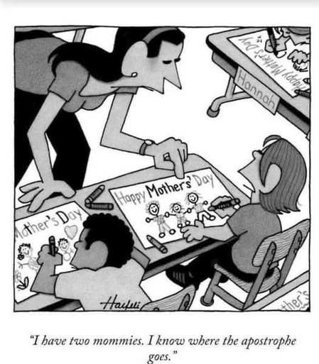 A cartoon shows a classroom where children are making Mother’s Day cards. A teacher bends over a student’s desk, pointing at the card. The student, a young girl with short hair, looks at the teacher and says, “I have two mommies. I know where the apostrophe goes.” Her drawing shows two women and children holding hands. The card says “Happy Mothers’ Day” with the apostrophe after the “s”. Other kids’ cards say “Mother’s Day” with the apostrophe before the “s”. A name tag on one desk reads “Hannah”. The classroom has small desks, chairs, and art supplies. The cartoon is in black and white and drawn in a simple, comic style.