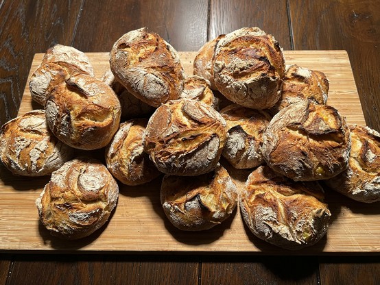 Auf einem dunklen Holztisch liegt ein Holzbrett. Darauf liegen zahlreiche frisch gebackene Brötchen mit rustikalem Ausbund.