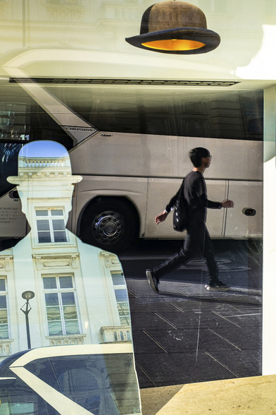Reflection shot in a shop window at the Magritte Museum in Brussels. On the left of the image a mirror in the shape of a man with a bowler hat reflects a classic building while a man dressed in black is passing by, a real bowler hat floating in the window above his head.