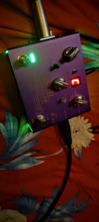 The photograph shows a purple pedal labeled "JOYO VOCAL LAB" placed on a red fabric with a floral pattern. The pedal has several controls, including four knobs and two footswitches, with two green LEDs  and a red LED display in the center. The knobs are silver with green highlights, and the footswitches are stainless steel. The pedal is connected to a power cable, and there is a metal XLR out jack at the left, also a metal XLR input Jack at the right. The background features a red fabric with blue and white floral designs
