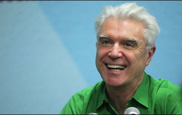 Retrato David Byrne con cabello corto y canoso peinado hacia atrás, sonriendo ampliamente. Tiene la piel clara y arrugas visibles alrededor de los ojos y la boca, lo que sugiere una expresión alegre y genuina. Lleva una camisa de color verde brillante y se aprecian dos micrófonos parcialmente visibles en la parte inferior de la imagen. El fondo es de un tono azul claro y difuminado.