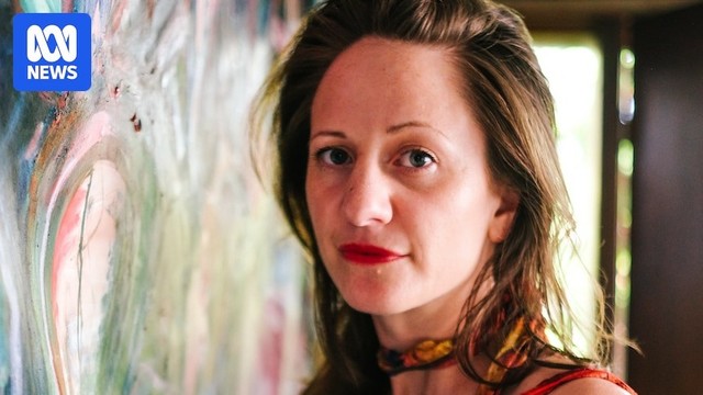 Photo of artist Nara Walker standing close to a painting and looking at the camera. She has long brown hair and red lipstick
