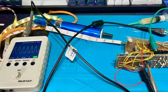 Photo of circuit board spaghetti with an attached oszilloscope that shows a sinus waveform. There is a blue silicone soldering desk protector underneath. LED strips are on the side glowing yellow.