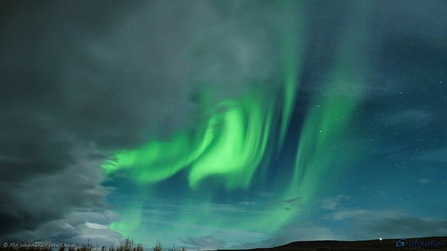 A colour photo of a night sky. From top centre to lower left the stars are hidden by a curtain of grey cloud. Behind, bright green aurora is folding and waving across a starry deep blue sky. At the bottom there's a horizon of trees and a hillside in silhouette.