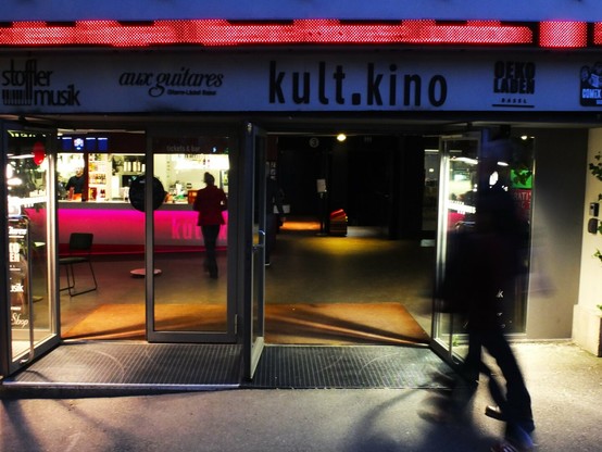 The image shows the entrance to a venue called "kult.kino" with glass doors open to reveal the interior. A red LED display runs along the top of the façade. Inside, there's a bar area with pink/red lighting and someone standing near it. The entrance has rubber floor mats at the doorway. To the left of the main entrance are signs for "stoffer musik" and "aux guitares," while to the right is what appears to be "DEKO BASEL." A silhouette of a person walking past the entrance is visible on the right side, casting a shadow on the pavement. The scene is captured at night, with the illuminated interior contrasting with the darker exterior.