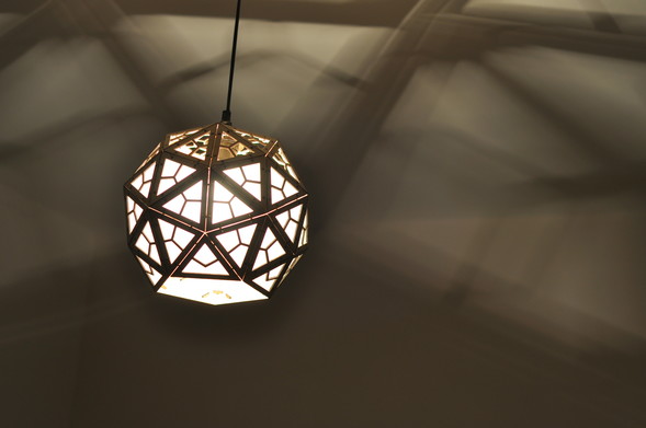 A picture of a lampshade set on a ceiling lamp in a dark room. The light is on.
The shade is made of laser cut wood and shape like a pentakis dodecahedron (like a regular dodecahedron with pentagonal faces, but those faces are replaced by small 5 faces pyramid).
The faces have a difuser except for the ones on top projecting geometrical shape of light on the ceilling.