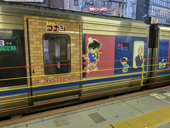 Sideview of a train, themed with detective conan anime elements and characters. There's 5 rubber ropes in front of it in red and yellow, stopping people from coming too close to the train while it's moving.