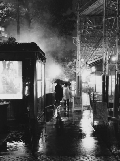 The image is in black and white and depicts a rainy urban street scene at night. There are people walking under umbrellas, wet pavements reflecting light, and structures like scaffolding or an awning. There's also a brightly lit billboard visible, contributing to the moody and atmospheric setting.