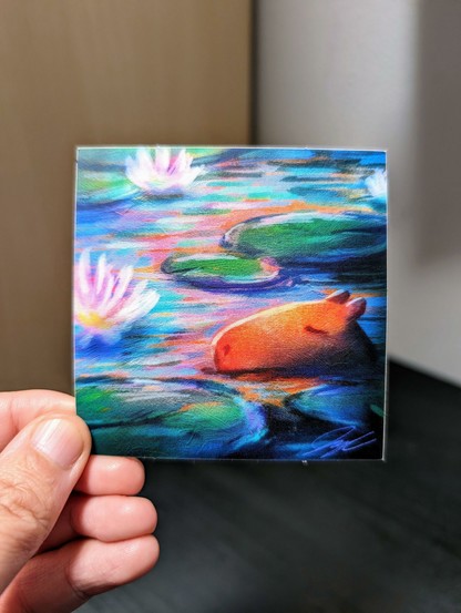 Photo of a left hand holding a square sticker. The sticker is a matte painting of a sleeping capybara among water lilies.