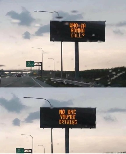 Digital road sign on a highway reads, “Who ya gonna call?” followed by, “No one. You’re driving.” Clouds and light rain in the background.