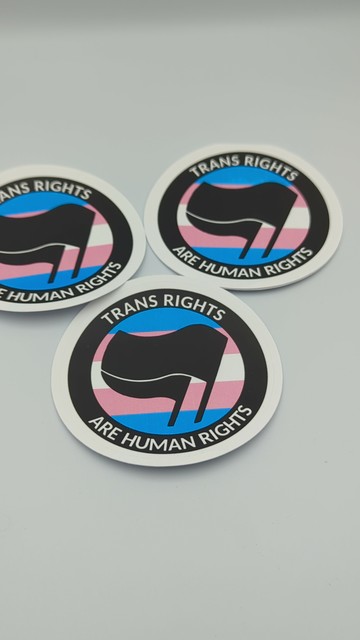 A photo of three 3 inch round stickers against a white backdrop. The design is a black ring with the words "trans rights are human rights" in sanserif capitals. In the centre of the ring is a double black flag, and a trans pride flags blue, pink and white bars fill the background.