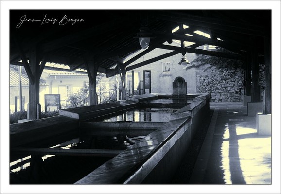 The image captures a serene and somewhat nostalgic scene of an old, covered washhouse or water basin area. The black-and-white color scheme adds a timeless, vintage feel to the photograph, emphasizing the textures and contrasts in the architectural elements and water reflections.
The washhouse features a long, rectangular water basin extending from the foreground to the background, with clean, calm water rippling slightly on the surface. The structure is supported by sturdy wooden beams, creating a shaded walkway alongside the basin. Light filters through openings in the roof and walls, casting patterned shadows that add depth to the scene.
In the background, a stone building with small windows complements the rustic and historical atmosphere, hinting at a rural or small-town setting. The overall composition, with its balance of light and shadow and the reflective water, suggests a place that was once a practical community gathering spot now captured in quiet stillness.
This setting carries cultural significance in many European regions where communal washhouses were essential social and functional spaces before modern plumbing became widespread. The image evokes a sense of heritage, tradition, and a slower pace of life, immortalized through the artistic choice of monochrome photography.