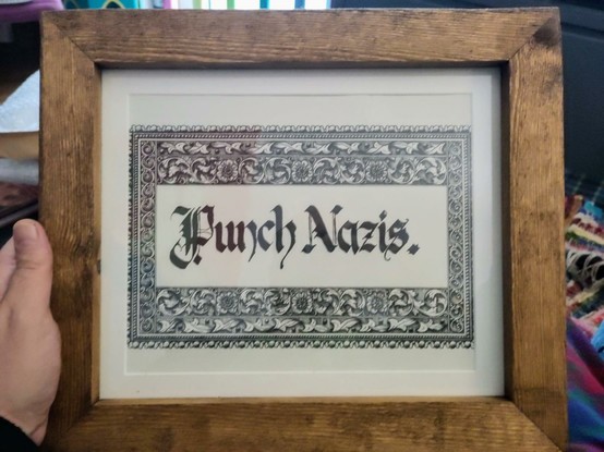 A calligraphy print with an intricate border is in a wooden frame. 
Inside the intricate border is the text "Punch Nazis." in calligraphy script.