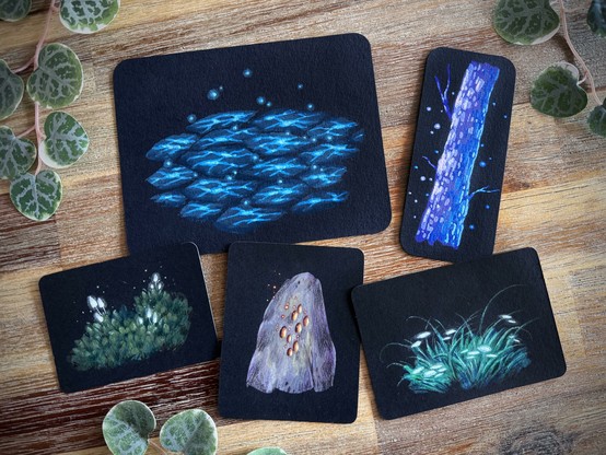 A photo of five small, black paper paintings with rounded corners placed on a wooden table and surrounded by leaves for decoration. One features glowing blue stone tiles, one features a ghostly pale tree, two feature greenery with glowing white plants/funghi, and the last one a standing stone with embers emanating from its core.