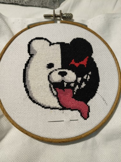a cross stitch of the character Monokuma from the Danganronpa games. He is a big teddybear with a normal white half and an evil black half with sharp teeth and a jagged glowing eye. his very long tongue is sticking out.
