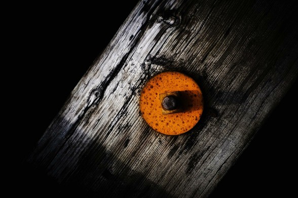 An orange washer is attached to a railway tie on a bridge. The background is pure black with only the grey weathered wood of the tie and the orange washer.