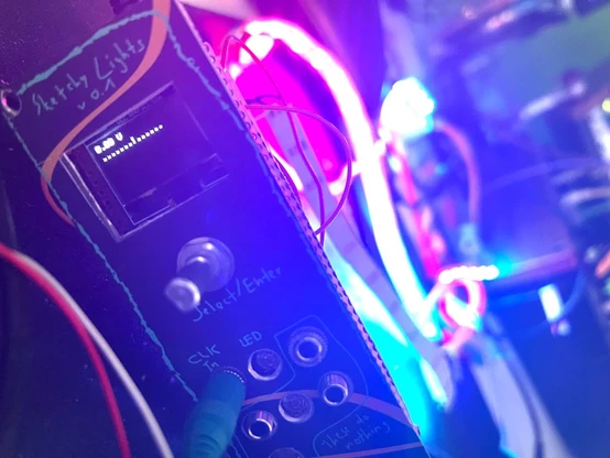 Photo of a DIY eurorack module with a lit screen and one cable connected. The screen shows dots for low level voltage and a bar for high level. One bar is shown. In the background a colourful LED strip is overexposing the image.