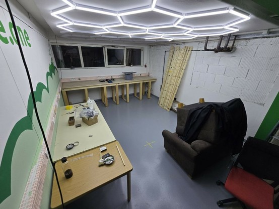 Ein Bild von dem neuen 4. Raum im vspace.one. Der Boden ist Grau und von der Decke beleuchtet ein sechseckiges Lichtmuster den Raum. Die Wände sind Weiß und auf der linken Wand ist zusätzlich eine grüne Rakete mit dessen Abgasen als Wolkenmuster aufgemalt. Außerdem lässt sich ein ebenfalls grüner vspace.one Schriftzug erahnen. Hinten befindet sich unter einem Kabelkanal mit Steckdosen und Netzwerkdosen eine Arbeitsplatte aus Holz mit einer Eurobox und Filamentrollen drauf. Darüber ist eine Reihe an Fenstern. Rechts an der Wand, bei der die Backsteine sichtbar sind, sind Holzbalken angelehnt und es sind Leitungen zu sehen. Links vorne ist ein Tisch mit einer beigen Platte, auf dem sich verschiedene Werkzeuge befinden. Rechts liegen zwei braune Sessel aufeinander. Davor steht ein Drehstuhl.