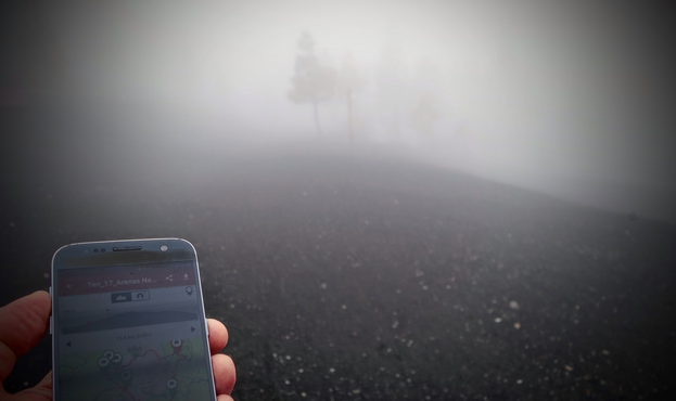 Wolken hüllen einen Berg in Nebel. Im Vordergrund hält eine Hand ein Smartphone mit einer GPS-Wanderkarte. In der Umgebung sind schwarze Vulkanasche und ein paar Bäume nur schemenhaft im Dunst zu erahnen.
