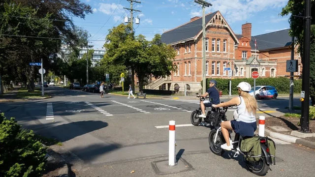 A view from Beacon Hill Park looking towards the Michigan Street advisory bike lanes. Two people on bicycle are crossing the street using the multi-use crossing with South Park School in the background.