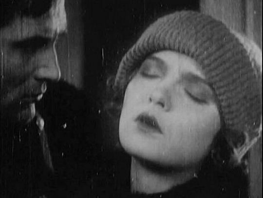 There is a man and a woman. The woman is wearing a hat, her eyes are closed and her dark hair is only partially visible. The man has short, dark hair and is looking at the woman.