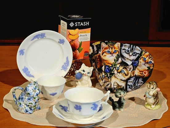 Tea tableau of playful ceramic kittens from the 1940s and 1970s, and a cat shaped Aynsley China creamer. There is vintage white china with blue Wedgwood styling, and a kitten patterned tea cozy. The tea is Stash peach black tea.