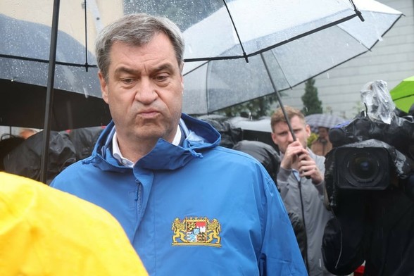 Prominenter Ministerpräsident der unter einem Regenschirm stehend die Backen aufbläst.[Quelle: https://www.tag24.de/nachrichten/politik/deutschland/politiker/markus-soeder/40-000-kraefte-in-hochwasser-region-in-bayern-im-einsatz-soeder-dankt-helfern-vor-ort-3204857]