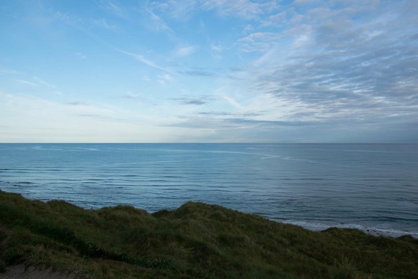 The image shows a scenic view of the coast, capturing the tranquil expanse of the ocean under a sky with scattered clouds. In the foreground, there are grass-covered sand dunes, which add a natural ruggedness to the scene. The overall mood is peaceful and expansive, typical of a coastal landscape.
