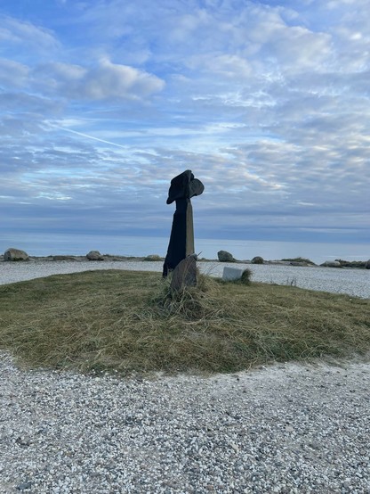 The image features a sculpture set in a coastal environment, likely by a beach or seaside. The sculpture appears abstract, consisting of a dark, textured column topped with a form that might suggest a head or an abstract shape. The ground surrounding the sculpture is covered with small stones and patches of grass. In the background, there is a calm sea and a scattered array of clouds in the sky, contributing to a serene and scenic view. The overall atmosphere is peaceful, enhanced by the natural light and soft color palette.