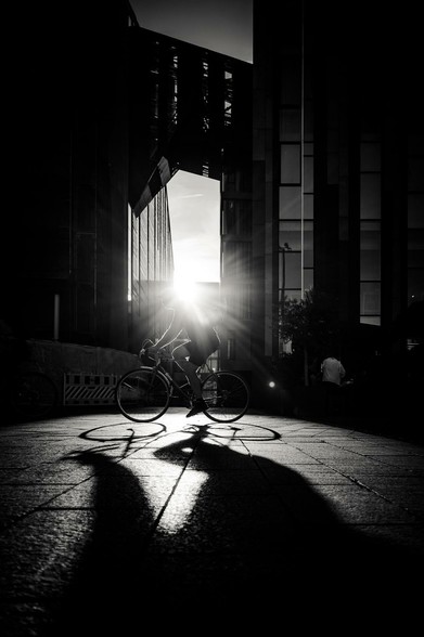 A lone cyclist navigates the stark, sun-drenched urban landscape. The harsh sunlight illuminates the person, casting long shadows across the pavement. Modern buildings loom in the background, their geometric shapes contrasting with the organic form of the cyclist and the bicycle.