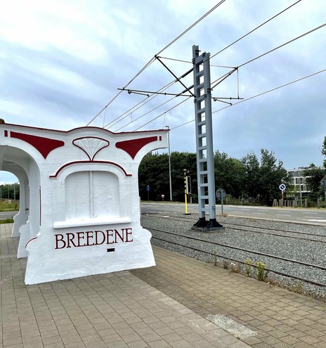This image shows a tram station named "Bredene." The station has a distinctive architectural style featuring white walls with red detailing. The structure appears to be part of the station design, likely for aesthetic or shelter purposes. There are multiple railway tracks visible, along with electrical lines above for the tram system. The backdrop includes a view of a road and some greenery, hinting at a suburban or semi-urban environment.