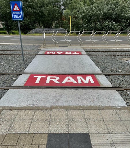 The image shows a tramway crossing integrated with pedestrian pathways. There's a road sign saying "VOORRANG TRAM," which translates to "Priority to Tram" in English, indicating that trams have the right of way at this crossing. The ground features text "TRAM" painted in red on a white concrete surface, likely to alert road users of the tram tracks. The pavement includes textured tiles for safety and accessibility, likely aiding visually impaired pedestrians. Additionally, there are metal railings on one side, probably to guide pedestrian movement and ensure safety near the tram tracks.