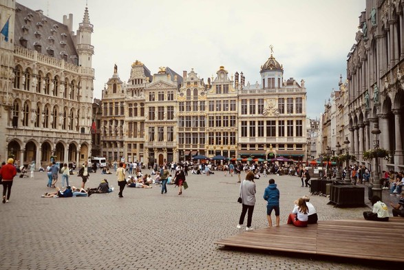 This image shows a bustling scene of the Grand Place (Grote Markt) in Brussels, Belgium. It's a famous historic square in the heart of Brussels, surrounded by ornate guild houses, the city's Town Hall, and the King's House or Breadhouse. The architecture prominently features Baroque and Gothic styles with gold trim and intricate details.

People are seen enjoying the atmosphere, with some sitting on the ground, walking, or chatting in groups. The sky is partly cloudy, suggesting a cool or mild weather. The cobbled square adds to the old-world charm of the setting. Overall, it's a lively and picturesque public space, typical of European historic centers.