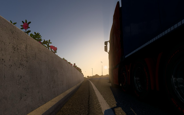 A low-angle view of a road with a concrete barrier on the left side, adorned with pink flowers. On the right side, a large truck is partially visible, with the sun setting in the background, casting a warm glow over the scene. The road stretches into the distance, lined with streetlights and trees.