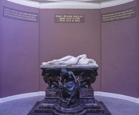 The image depicts a tomb or memorial of Percy Bysshe Shelley, a famous English poet. The memorial features a large, sculpted figure lying on a stone sarcophagus. The tomb is intricately designed, with artistic details surrounding the central figure. The walls have inscriptions mentioning Shelley’s name, birthdate, and date of death, as well as poetic references. The overall ambiance suggests a place of remembrance and reverence for the poet.

Image Credits: Wikimedia / Godot13 / CC BY-SA 4.0