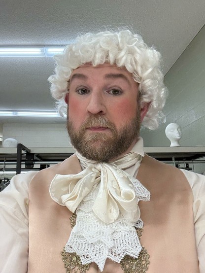 A person is dressed in 18th-century attire, featuring a white curly wig, a beige vest, and a decorative cravat. They display a whimsical expression, standing in a room with light-colored walls and mannequin heads in the background.